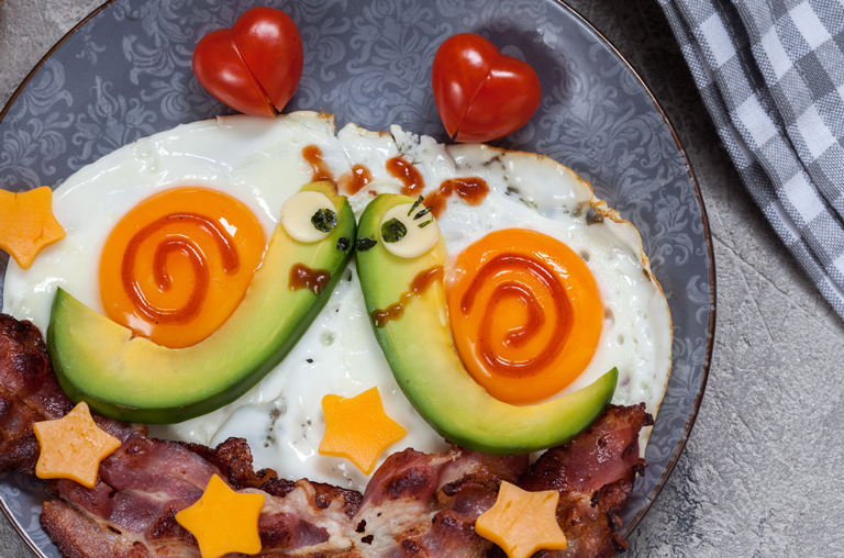 Abbildung Spiegelei mit Avocado, Bacon und Tomaten in Herzform. Avocados fiormiert als küssende Schnecken